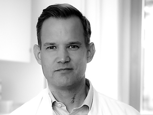 Black-and-white portrait photo of a middle-aged person of male appearance with a large forehead and protruding ears. They are wearing a white collared shirt.