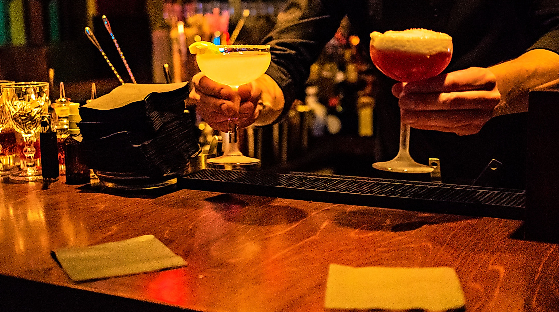 Zwei Cocktails werden an einer Bar bei warmem Licht serviert, daneben Gläser und Barutensilien
