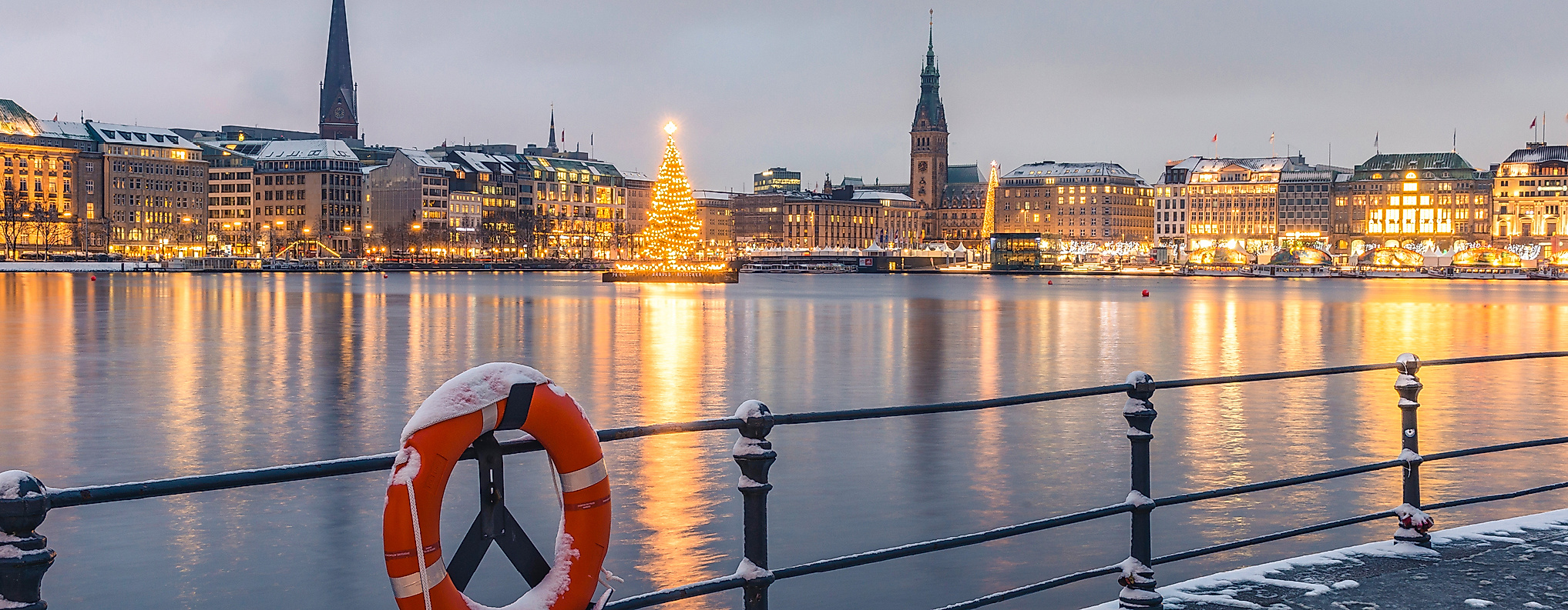 Winterliche Alstersilhouette mit Weihnachtsbaum, verschneitem Geländer und festlich beleuchtetem Jungfernstieg in Hamburg.