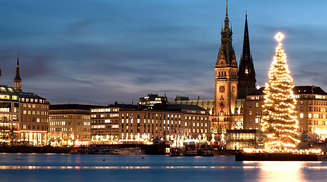 Beleuchteter Weihnachtsbaum auf der Alster vor dem Hamburger Rathaus und festlich erleuchteter Innenstadt bei Abenddämmerung.
