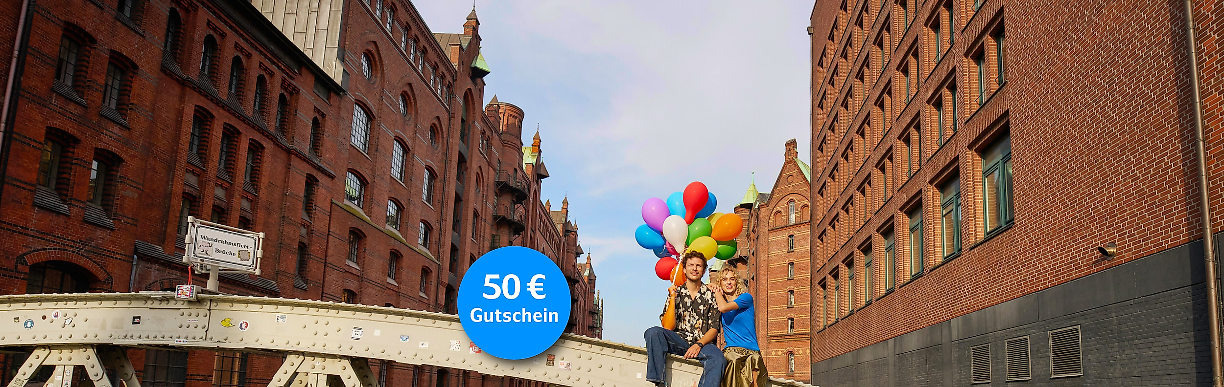 Mann und Frau sitzen auf einer Brücke mit Luftballons in der Hand und der Speicherstadt im Hintergrund