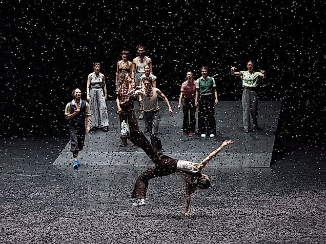 A group of dancers stands on a sloped stage while, in the foreground, one person balances impressively on one hand as confetti swirls through the air.