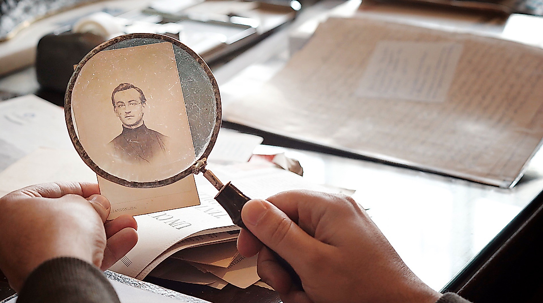 Hände halten eine Lupe über ein altes, vergilbtes Foto auf einem Schreibtisch mit weiteren Papieren und Dokumenten.
