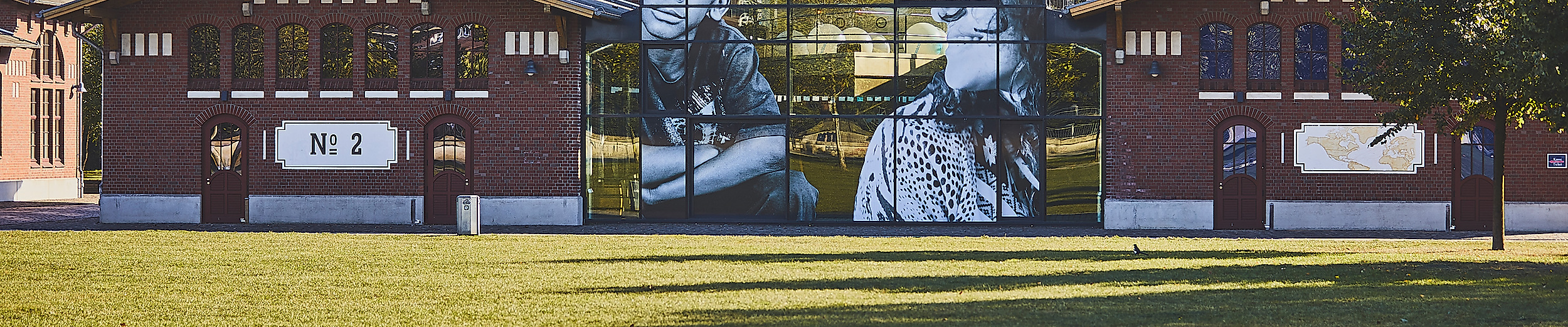 Backsteingebäude des Auswanderermuseums BallinStadt mit Glasfassade und Porträtmotiven, grüne Wiese im Vordergrund
