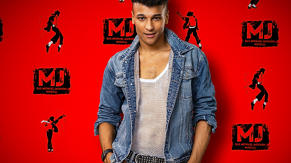 Musicaldarsteller Prince Damien in Jeansjacke vor Wand mit MJ - Das Michael Jackson-Logos