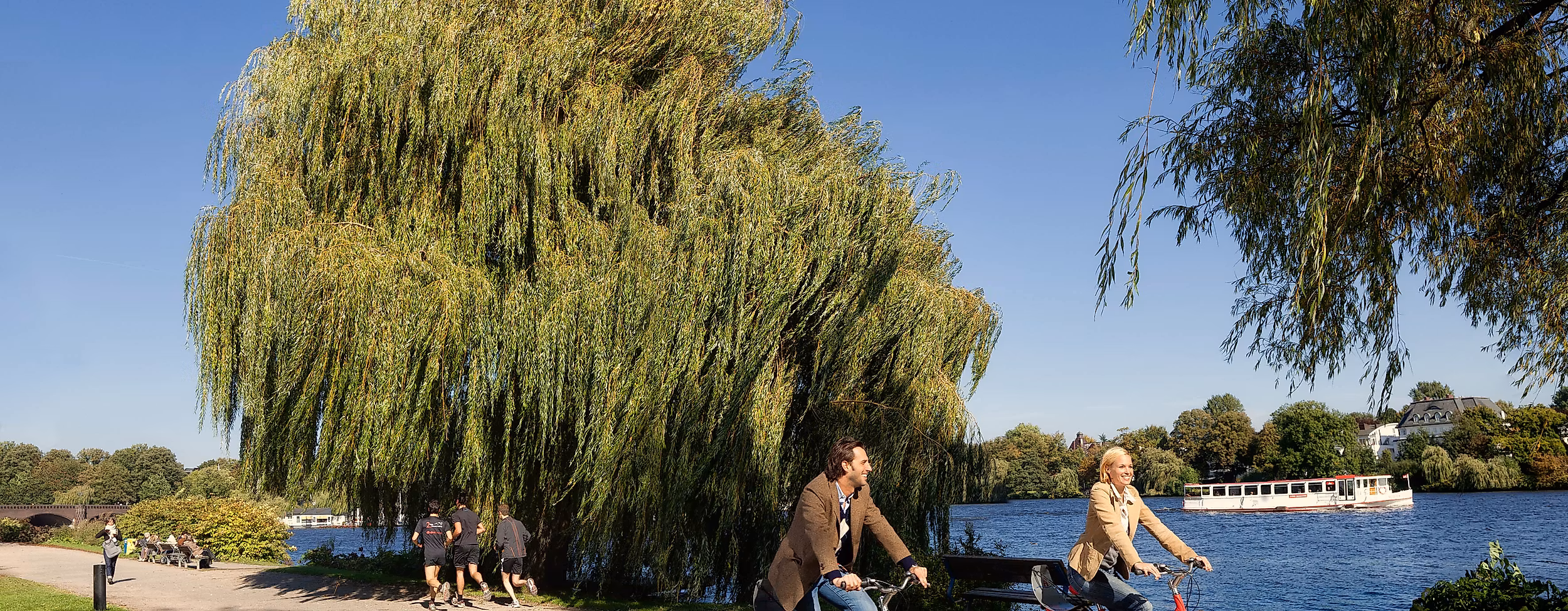 Zwei Personen radeln mit Stadträdern am Ufer der Alster entlang unter blauem Himmel.