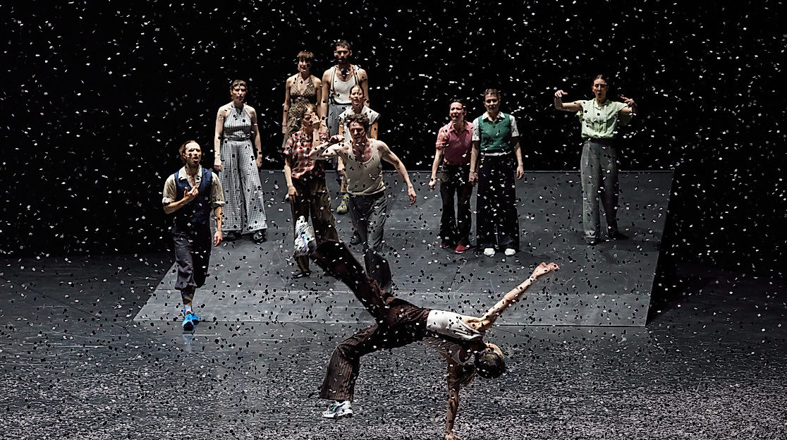 A group of dancers stands on a sloped stage while, in the foreground, one person balances impressively on one hand as confetti swirls through the air.
