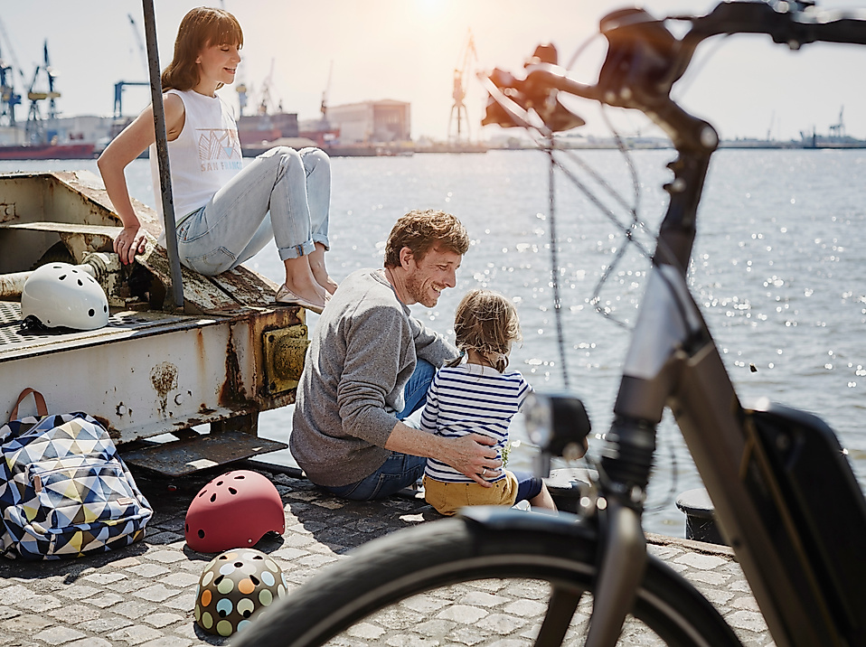 Eine Familie mit Kind sitzt an der Hafenkante, im Vordergrund ist ein Fahrrad zu sehen