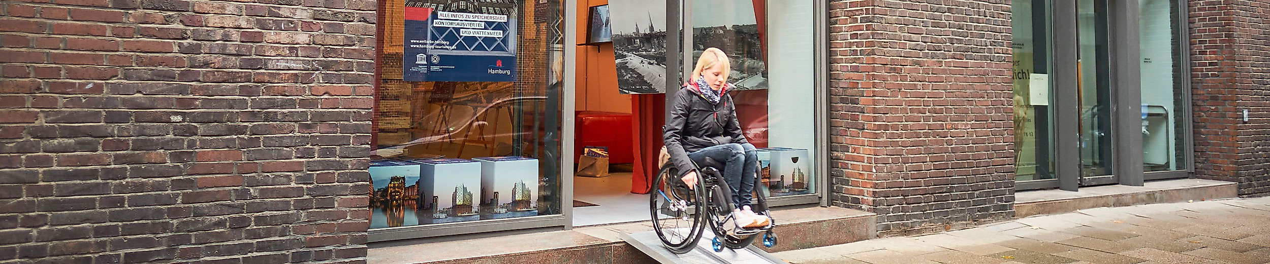 Rollstuhlfahrerin nutzt mobile Rampe am barrierefreien Eingang des Welterbe Info Points in Backsteingebäude.