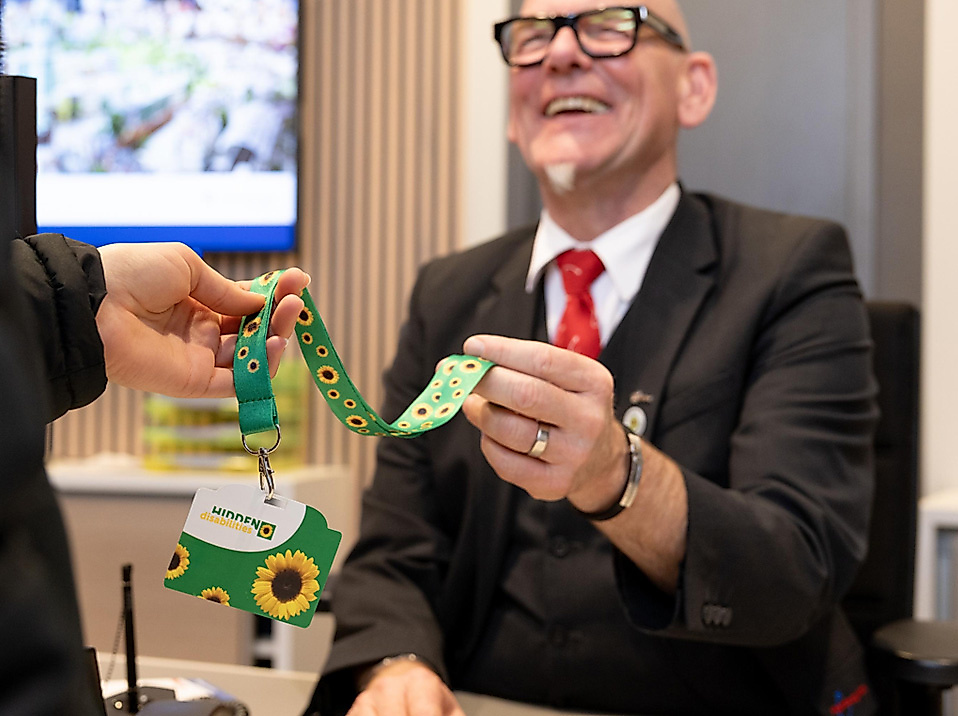Mann nimmt Umhängeband entgegen, auf dem sich das Sunflower-Symbol für nicht-sichtbare Behinderungen befindet
