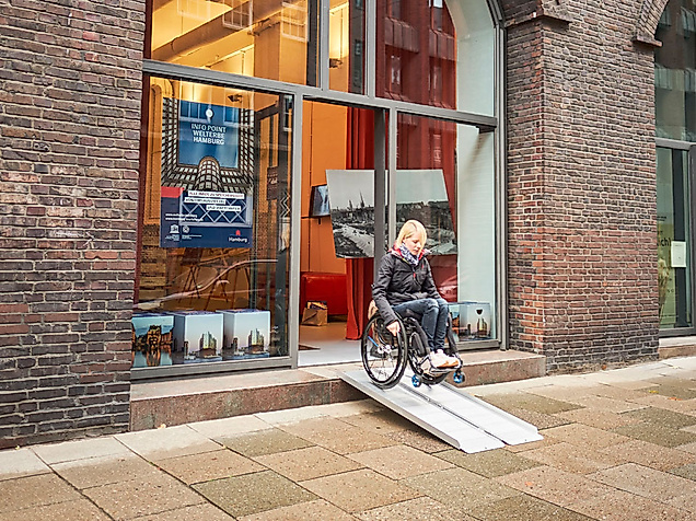 Rollstuhlfahrerin nutzt mobile Rampe am barrierefreien Eingang des Welterbe Info Points in Backsteingebäude.