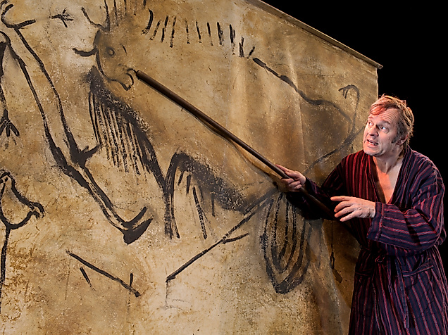 Szenenbild Cavemen: Ein Mann in Bademantel zeigt mit einem Stock auf eine große Wand mit Zeichnungen
