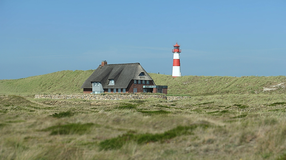Reetdachhaus vor Dünenlandschaft mit rot-weißem Leuchtturm bei klarem Himmel.
