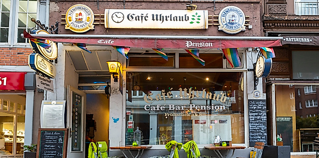 Außenansicht des Café Urlaub in Hamburg-St. Georg mit bunter Deko, Klappstühlen und einladender Fensterfront.