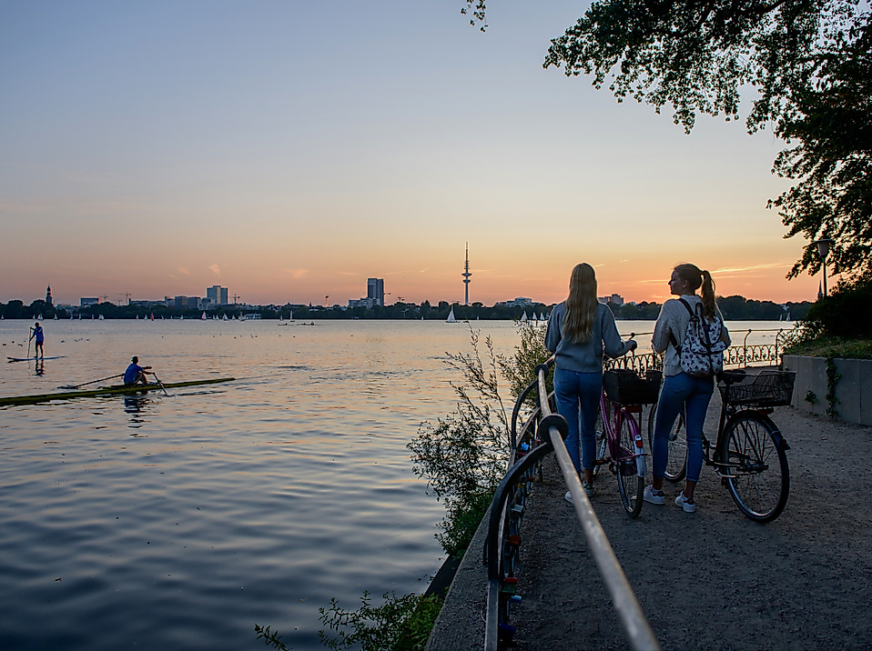 Zwei Radfahrer:innen blicken bei Sonnenuntergang über die Binnenalster mit Kajakfahrern und Stadtsilhouette.