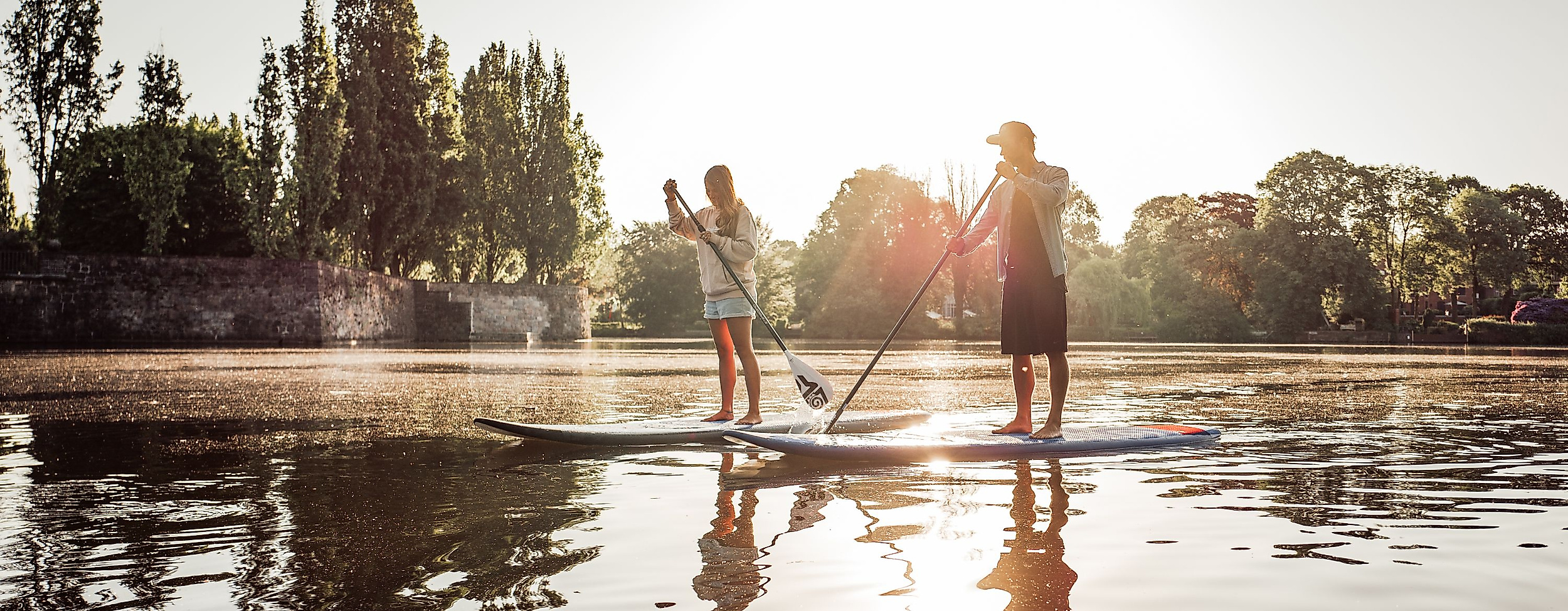 Zwei Personen beim Stand-up-Paddling im Gegenlicht auf ruhigem Wasser vor Bäumen und Abendsonne