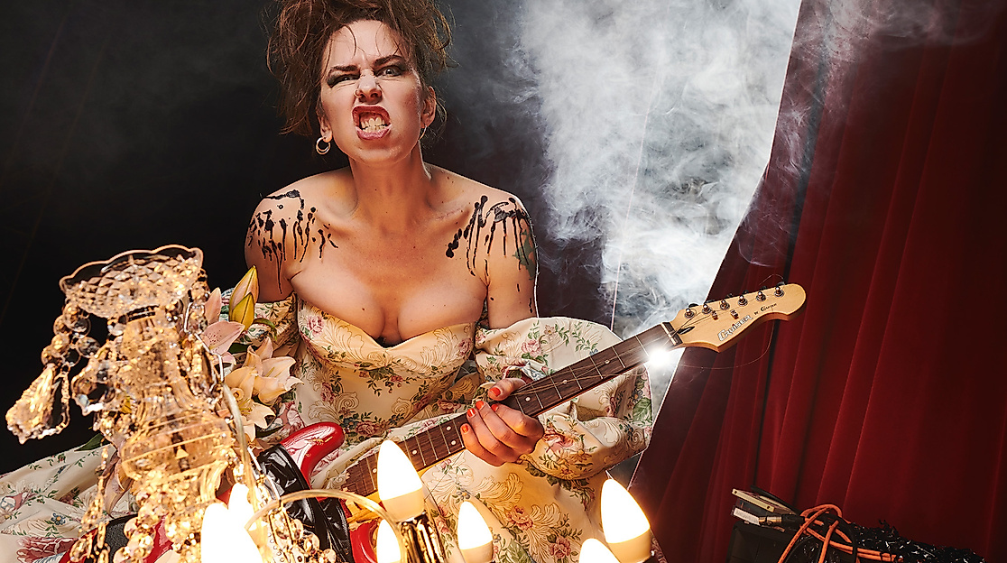 Emma Frankland, eine weiblich gelesene Person mit braunen Haaren steht mit einer E-Gitarre und rockigem Gesichtsausdruck vor einem Vorhang. Im Hintergrund ist Rauch zu sehen, während im Vordergrund ein Kronleuchter ist.