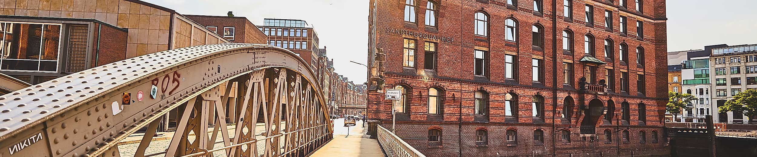 Ein sonniger Tag in der Hamburger Speicherstadt. Im Bildvordergrund führt eine historische Stahlbrücke mit Holzbohlen auf ein imposantes rotes Backsteingebäude mit Türmchen und Giebeln zu. Die Sonne scheint durch die Gebäudekante hindurch und erzeugt eine warme Lichtstimmung.