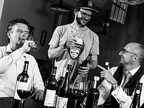 Slanted black-and-white photo of three people, one dressed casually and two more formally, in front of a table full of wine glasses and bottles. One person is drinking and the other two are looking at each other and smiling, both also holding wine glasses