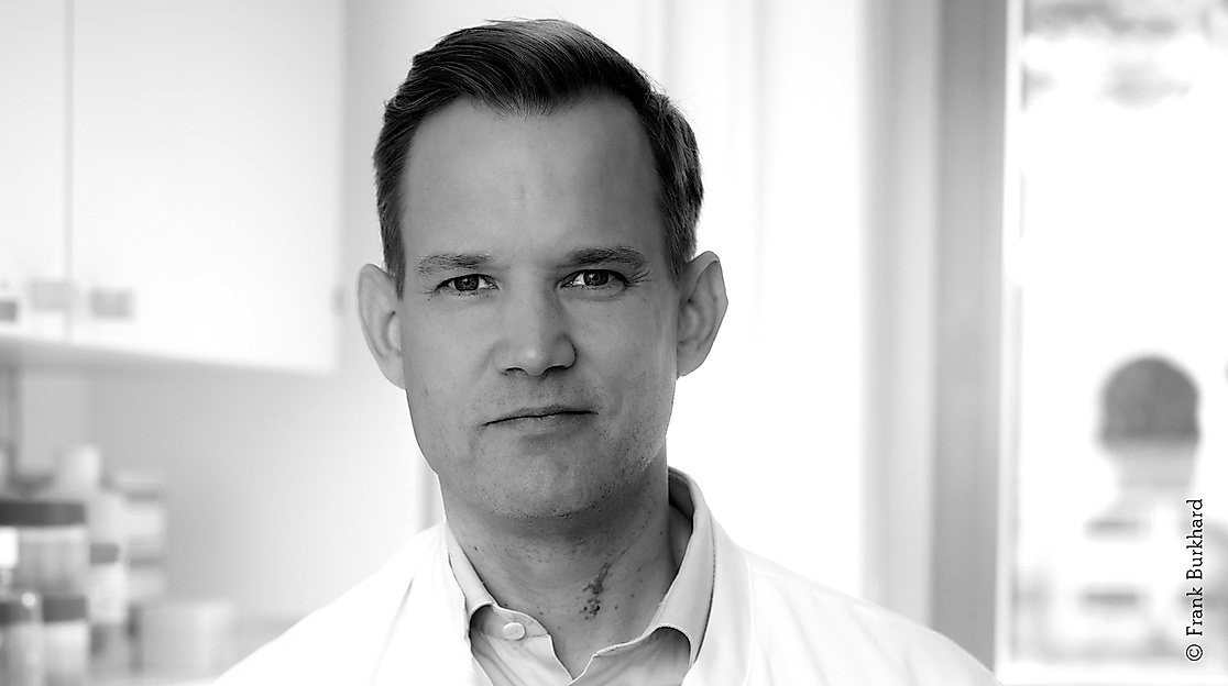 Black-and-white portrait photo of a middle-aged person of male appearance with a large forehead and protruding ears. They are wearing a white collared shirt.