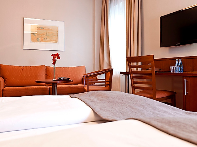 Helles Zimmer mit Doppelbett, braunem Sofa, Schreibtisch und großem Fernseher an der Wand.