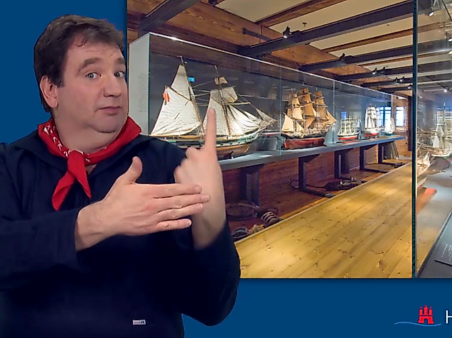 Man signing in International Sign in front of a maritime exhibition in Hamburg – accessible cultural information.