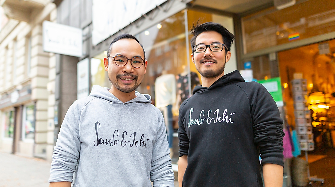 Zwei lächelnde Männer im Hoodie stehen auf dem Gehweg vor dem japanischen Restaurant Sando & Ichi in Hamburg-St. Georg, Blick in die Kamera 