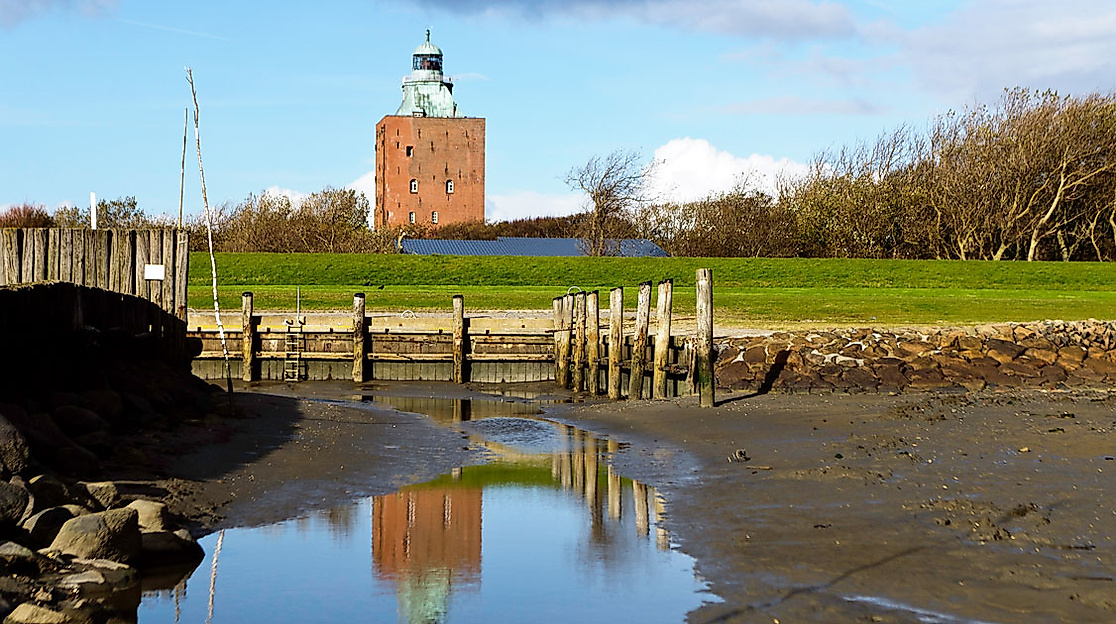 Leuchtturm Neuwerk hinter Deich und Holzsteg, Spiegelung im Schlick bei Ebbe unter blauem Himmel