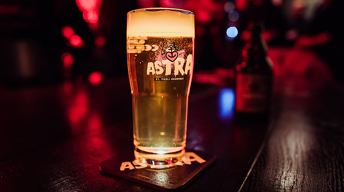 Stimmungsvoll beleuchtete Astra-Kneipe mit Holztischen, roten Sitzbänken und Blick auf den Zapfhahnbereich