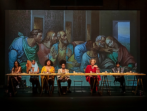 Performers sit at a long table in front of a projection of Da Vinci’s »Last Supper« — a contemporary staging with a diverse cast.