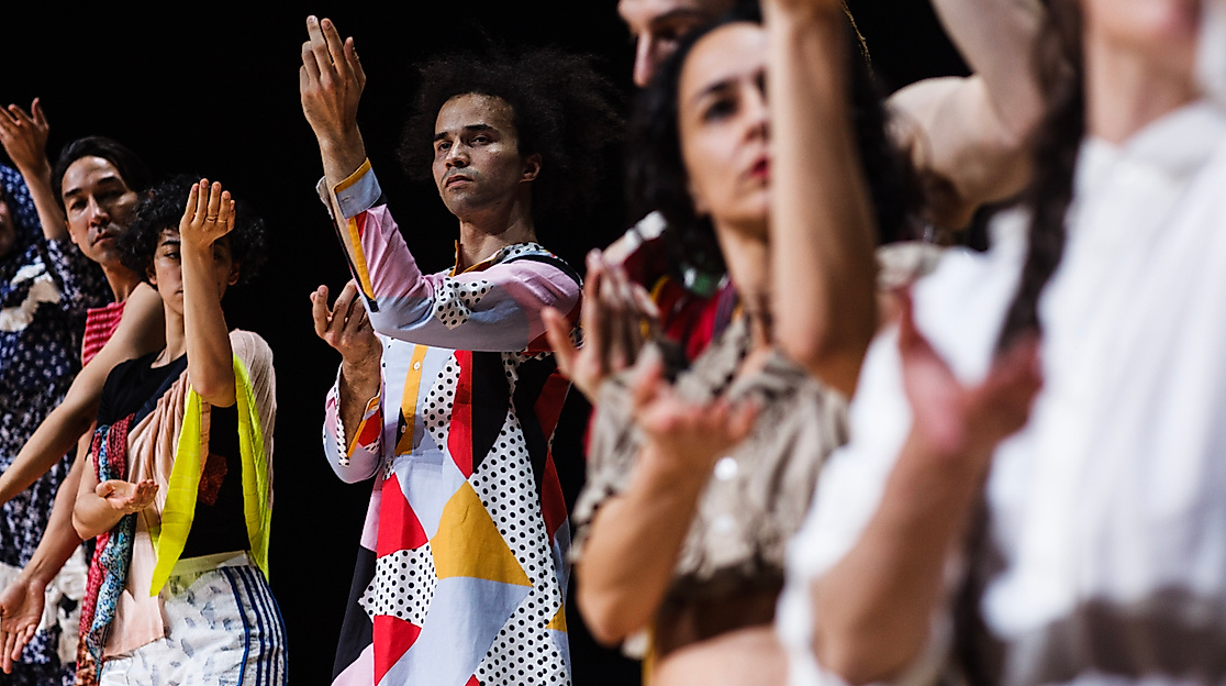 A group of dancers stand in a row and synchronously perform an expressive hand movement, focusing on one person in a colourful, geometrically patterned outfit.