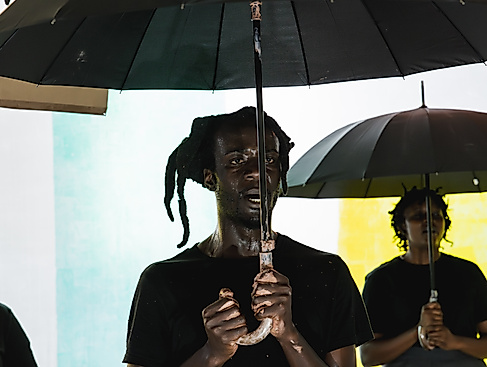 Excerpt from the production DAMBUDZO by nora chipaumire. A person stands holding an umbrella in front of a colorful wall. Behind them, two other people are visible in the background.