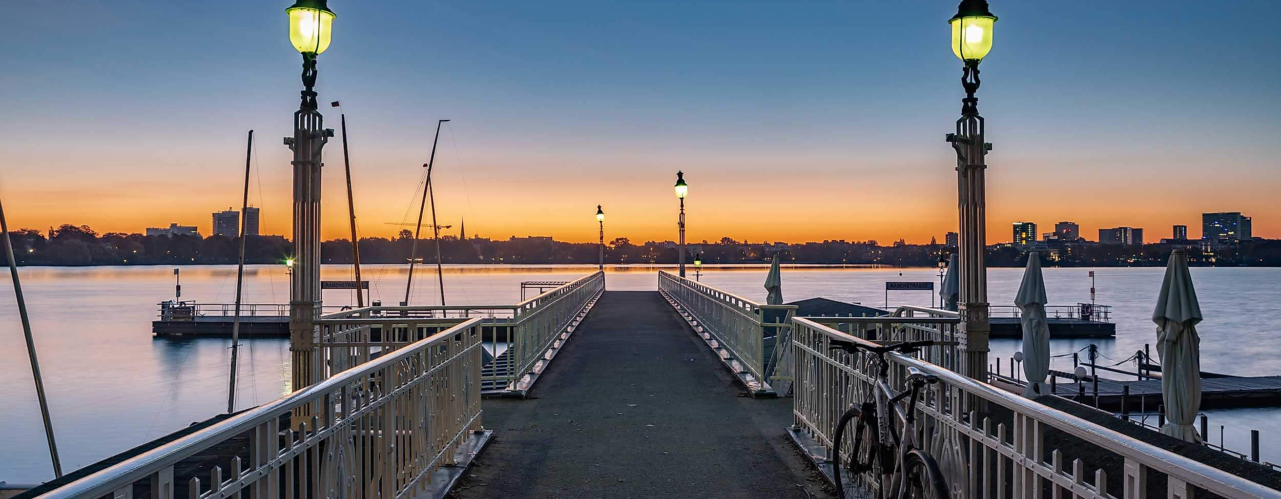 Steg mit Laternen und Fahrrädern an der Außenalster bei Sonnenuntergang mit Blick auf Hamburg