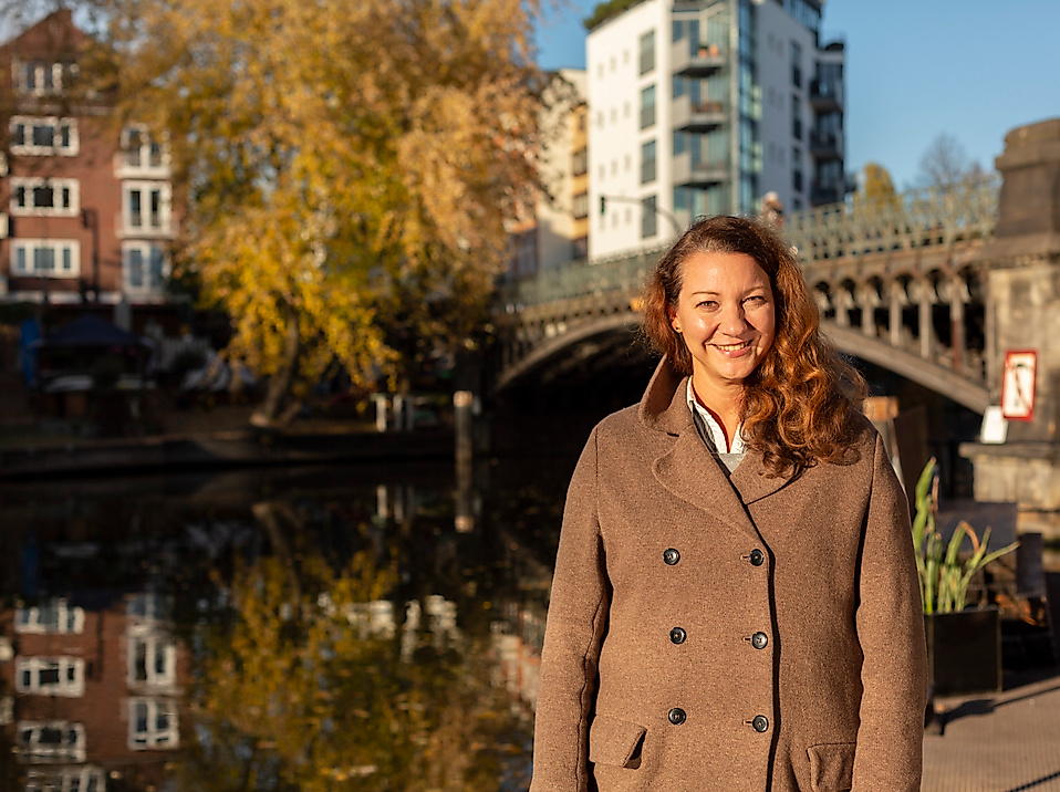Lächelnde Frau vor Brücke und Kanalufer an der Hamburger Alster bei Sonnenlicht und herbstlicher Stimmung