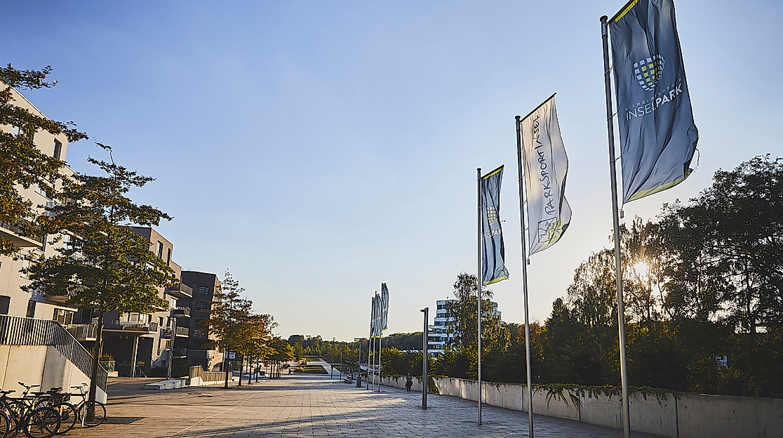 Promenade mit Fahnen im Wilhelmsburger Sommerpark bei Sonnenschein, umgeben von Bäumen und modernen Gebäuden