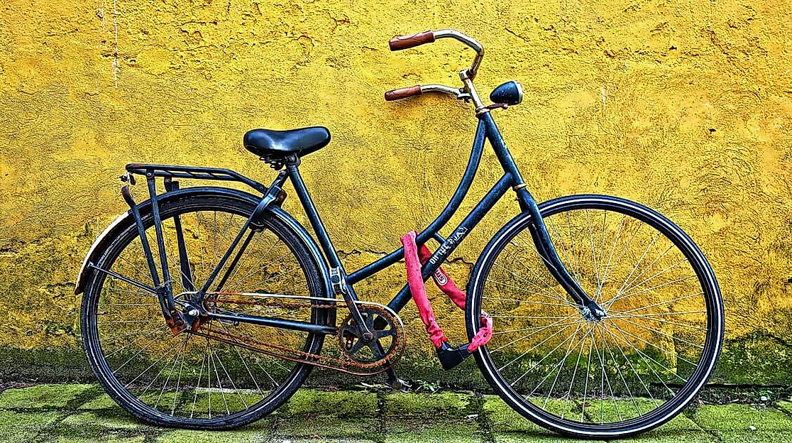 Angeschlossenes Fahrrad mit rotem Kettenschloss vor gelber Wand auf grünem Bodenbelag