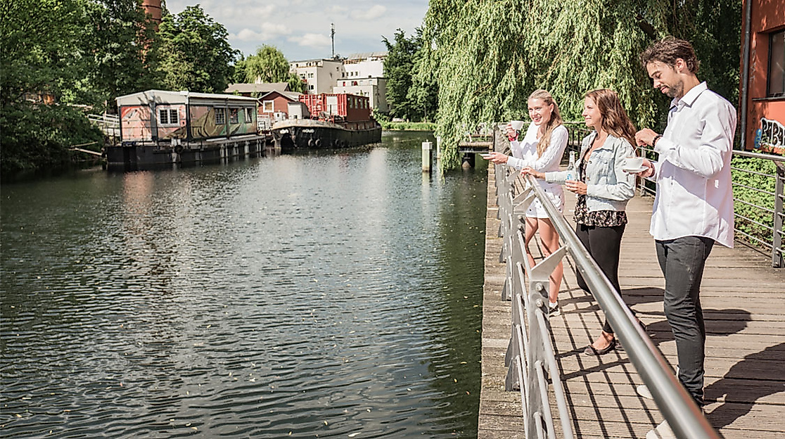 Menschen stehen an der Uferpromenade des Veringkanals in Wilhelmsburg, umgeben von Wasser, Bäumen und Hausbooten