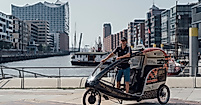 Rikscha-Fahrer mit Fahrgästen in der HafenCity, im Hintergrund die Elbphilharmonie und moderne Architektur.