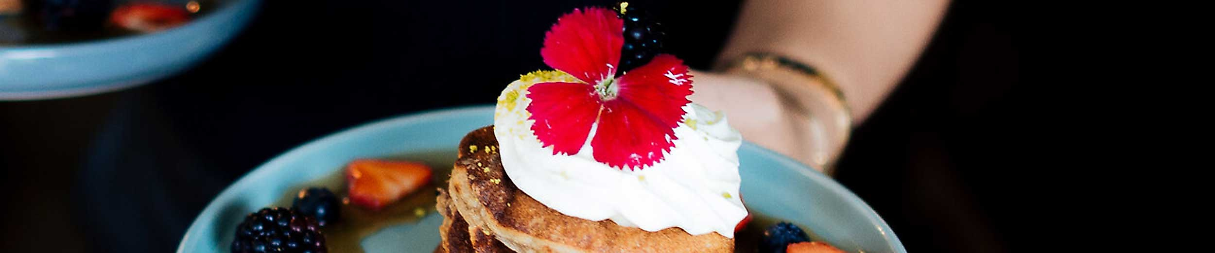 Ein kunstvoll angerichteter Stapel Pancakes auf einem blauen Teller, garniert mit Sahne, einer essbaren roten Blüte und Zesten von Limette. Drumherum frische Brombeeren und Erdbeerstücke. Eine Hand serviert das Frühstück – perfekte Szene für einen genussvollen Start in den Tag in einem Hamburger Café, das mit der Hamburg CARD Ermäßigungen bietet