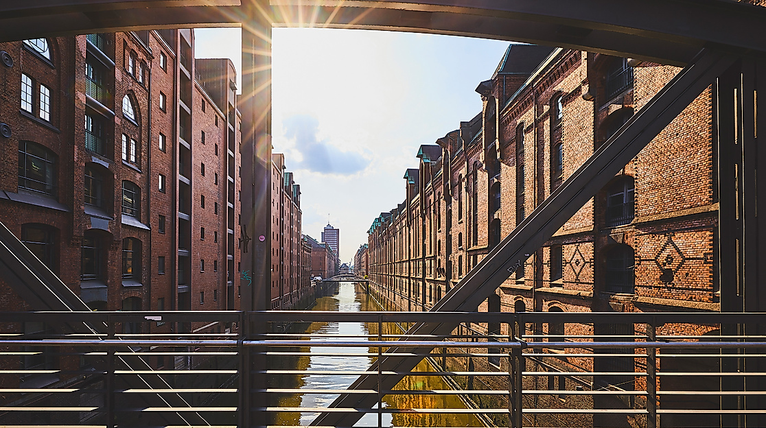 Blick durch eine Stahlbrücke auf die sonnenbeschienenen Backsteinfassaden der Speicherstadt entlang des Fleets