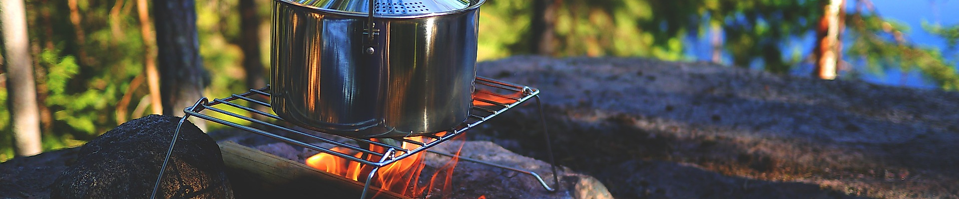 Kochtopf über offenem Feuer auf Holzunterlage im Wald nahe einem See beim Camping