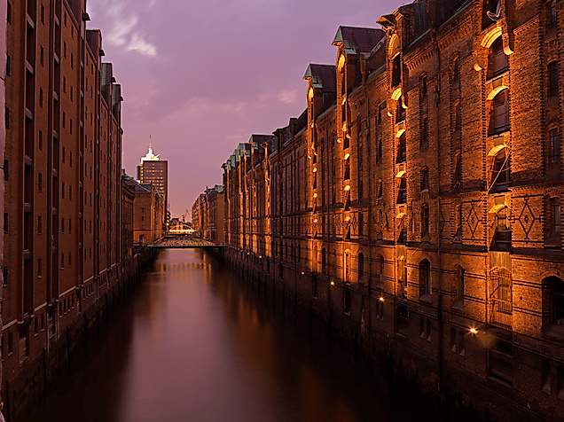 Beleuchtete Lagerhäuser der Speicherstadt spiegeln sich bei Abenddämmerung im ruhigen Fleet.