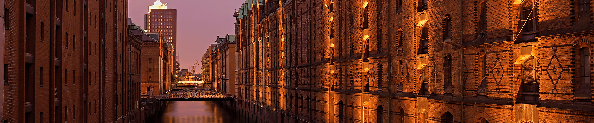 Beleuchtete Lagerhäuser der Speicherstadt spiegeln sich bei Abenddämmerung im ruhigen Fleet.