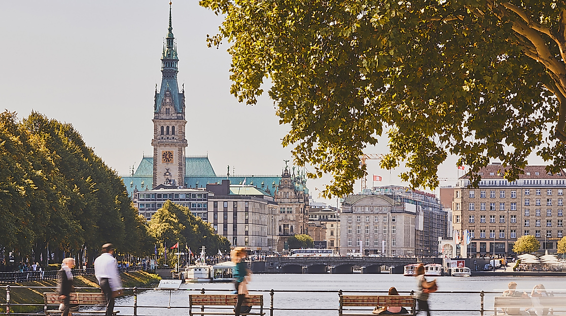 Blick über die Binnenalster auf das Hamburger Rathaus, eingerahmt von Bäumen und Sitzbänken
