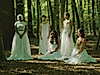 Five performers in long white dresses pose in the forest. Three of them are standing, two are sitting on the ground. None of them are looking at the camera.