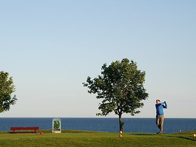 Golfer schlägt am Golfpark Fehmarn mit Blick auf die Ostsee ab, umgeben von Bäumen und Küstenlandschaft