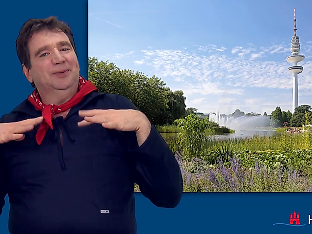  Man signing in International Sign in front of a green park landscape with Hamburg’s TV tower in the background.