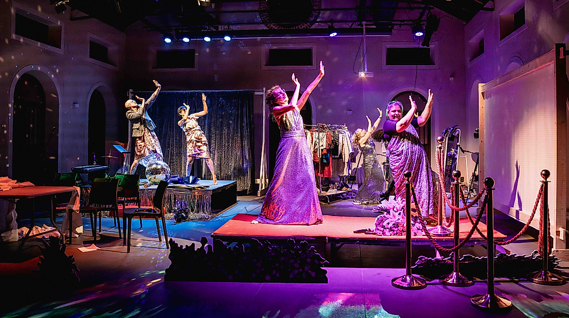 The performers dance around the stage to Bollywood music, making big gestures. There are colorful lights and the glow of a disco ball.