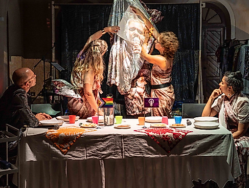 Zwei Performerinnen sitzen an den kurzen Seiten einer weißen Tafel und schauen auf ein Gruppe hinter der Tafel. Mit Stoffen und Tellern bilden diese eine Skulptur auf die Video projiziert wird.