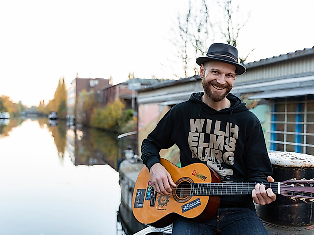 Mann mit Gitarre und Hut sitzt am Wasser vor Hausboot in Hamburg, lächelt in die Kamera bei Herbstlicht